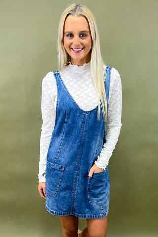 Woman wearing a blue denim dress over a white long-sleeve shirt against a green background