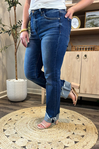Close-up of a woman wearing high-waisted dark blue straight-leg jeans with distressed, rolled cuffs. She pairs them with a white ribbed tank top and nude block-heel sandals, standing on a woven circular rug with a potted plant and wooden shelving in the background.