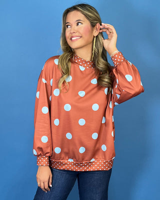 Rust polka dot long sleeve top with oversized light blue dots, contrasting mini dot mock neckline, cuffs and hem, and a relaxed fit.