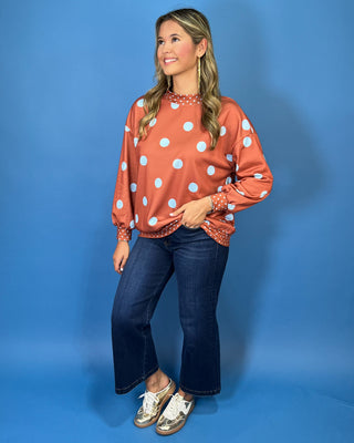 Rust polka dot long sleeve top with oversized light blue dots, contrasting mini dot mock neckline, cuffs and hem, and a relaxed fit.