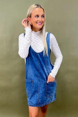 Woman wearing a denim dress over a white turtleneck sweater against a green background