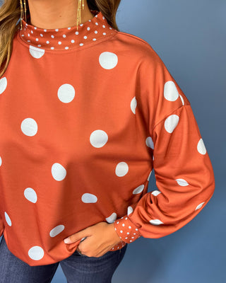 Rust polka dot long sleeve top with oversized light blue dots, contrasting mini dot mock neckline, cuffs and hem, and a relaxed fit.