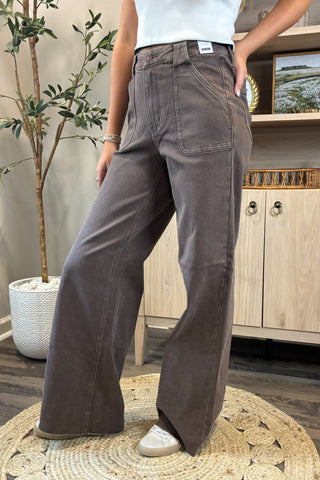 Close-up of a woman wearing high-waisted brown wide-leg jeans with large front patch pockets. She pairs them with a white sleeveless knit top and white sneakers, standing on a woven circular rug with a potted plant and wooden shelving in the background.