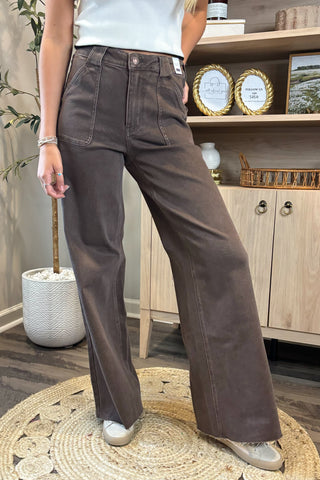 Close-up of a woman wearing high-waisted brown wide-leg jeans with large front patch pockets. She pairs them with a white sleeveless knit top and white sneakers, standing on a woven circular rug with a potted plant and wooden shelving in the background.