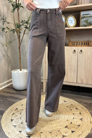 Close-up of a woman wearing high-waisted brown wide-leg jeans with large front patch pockets. She pairs them with a white sleeveless knit top and white sneakers, standing on a woven circular rug with a potted plant and wooden shelving in the background.