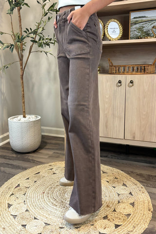 Close-up of a woman wearing high-waisted brown wide-leg jeans with large front patch pockets. She pairs them with a white sleeveless knit top and white sneakers, standing on a woven circular rug with a potted plant and wooden shelving in the background.