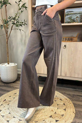 Close-up of a woman wearing high-waisted brown wide-leg jeans with large front patch pockets. She pairs them with a white sleeveless knit top and white sneakers, standing on a woven circular rug with a potted plant and wooden shelving in the background.