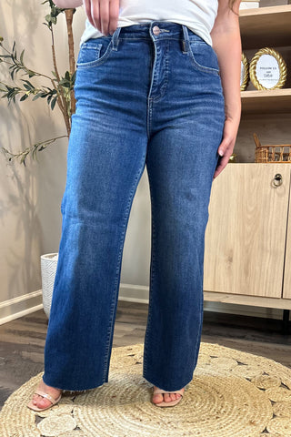 Close-up of a woman wearing high-waisted medium-wash wide-leg jeans. She pairs them with a white ribbed tank top and nude block-heel sandals, standing on a woven circular rug with a potted plant and wooden shelving in the background.