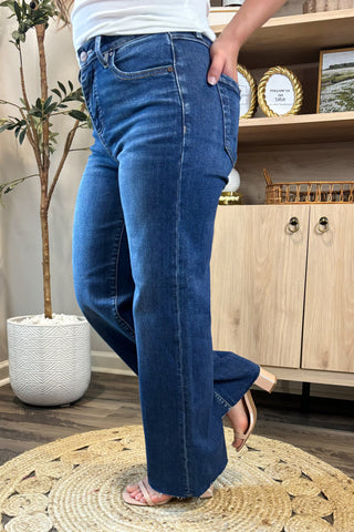 Close-up of a woman wearing high-waisted medium-wash wide-leg jeans. She pairs them with a white ribbed tank top and nude block-heel sandals, standing on a woven circular rug with a potted plant and wooden shelving in the background.