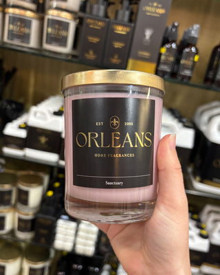 Hand holding a candle labeled 'Orleans Home Fragrances' in front of shelves with similar products.