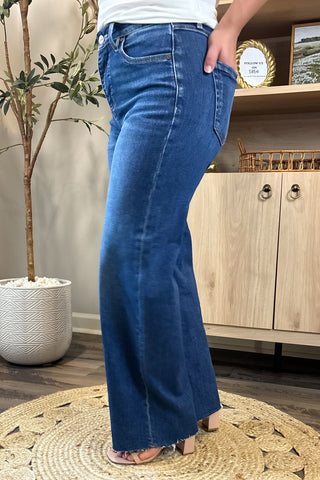 Close-up of a woman wearing high-waisted medium-wash wide-leg jeans. She pairs them with a white ribbed tank top and nude block-heel sandals, standing on a woven circular rug with a potted plant and wooden shelving in the background.