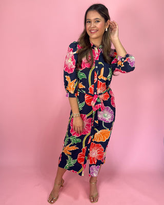Navy midi shirt dress with bold multicolor floral print, featuring a collared neckline, button-front closure, three-quarter sleeves, and a self-tie waist belt, styled with gold strappy heels against a pink background.