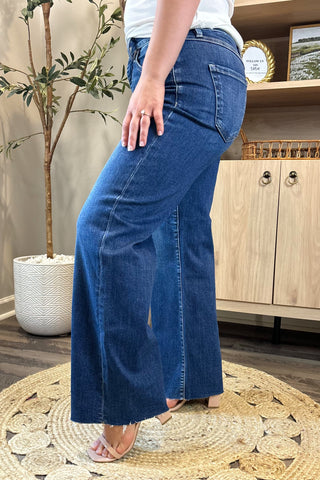 Close-up of a woman wearing high-waisted medium-wash wide-leg jeans. She pairs them with a white ribbed tank top and nude block-heel sandals, standing on a woven circular rug with a potted plant and wooden shelving in the background.