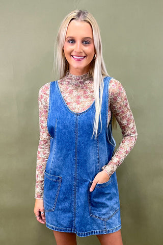 Woman wearing a blue denim pinafore dress over a floral long-sleeve shirt against a green background