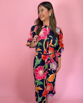 Navy midi shirt dress with bold multicolor floral print, featuring a collared neckline, button-front closure, three-quarter sleeves, and a self-tie waist belt, styled with gold strappy heels against a pink background.