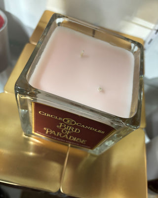 Square candle in a glass container with a label on a gold stand.