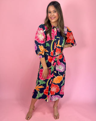 Navy midi shirt dress with bold multicolor floral print, featuring a collared neckline, button-front closure, three-quarter sleeves, and a self-tie waist belt, styled with gold strappy heels against a pink background.