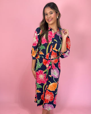 Navy midi shirt dress with bold multicolor floral print, featuring a collared neckline, button-front closure, three-quarter sleeves, and a self-tie waist belt, styled with gold strappy heels against a pink background.