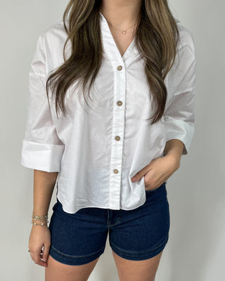 Smiling woman styled in a crisp white button-down with cuffed sleeves, dark wash frayed denim shorts, and clear slide sandals with simple gold jewelry.