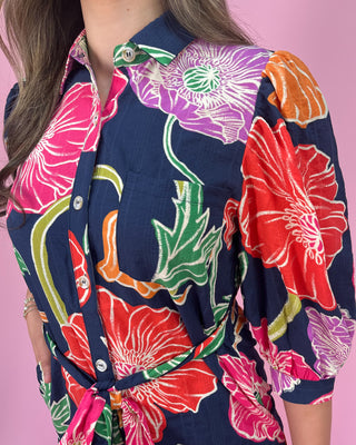 Navy midi shirt dress with bold multicolor floral print, featuring a collared neckline, button-front closure, three-quarter sleeves, and a self-tie waist belt, styled with gold strappy heels against a pink background.
