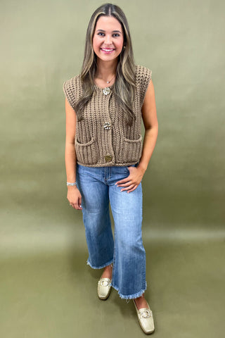 Woman wearing a brown knitted vest and blue jeans against a green background