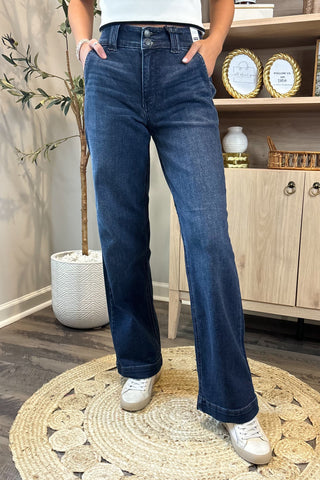 Close-up of a woman wearing high-waisted dark blue wide-leg jeans with a double-button closure. She pairs them with a white sleeveless knit top and white sneakers, standing on a woven circular rug with a potted plant and wooden shelving in the background.