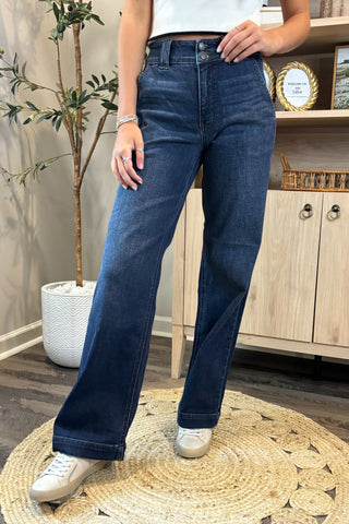 Close-up of a woman wearing high-waisted dark blue wide-leg jeans with a double-button closure. She pairs them with a white sleeveless knit top and white sneakers, standing on a woven circular rug with a potted plant and wooden shelving in the background.
