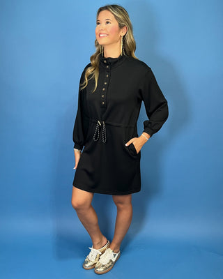Black long sleeve mini dress with a buttoned mock neckline, adjustable drawstring waist, side pockets, and an easy sporty-chic silhouette.