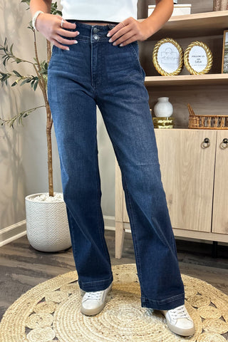 Close-up of a woman wearing high-waisted dark blue wide-leg jeans with a double-button closure. She pairs them with a white sleeveless knit top and white sneakers, standing on a woven circular rug with a potted plant and wooden shelving in the background.