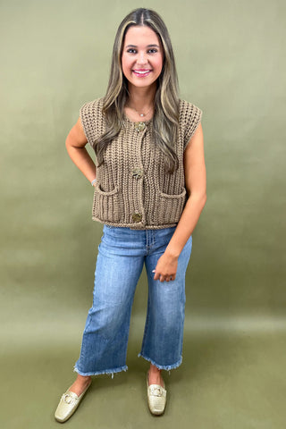 Woman wearing a brown knitted sleeveless sweater and blue jeans against a green background