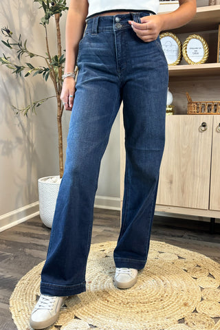 Close-up of a woman wearing high-waisted dark blue wide-leg jeans with a double-button closure. She pairs them with a white sleeveless knit top and white sneakers, standing on a woven circular rug with a potted plant and wooden shelving in the background.