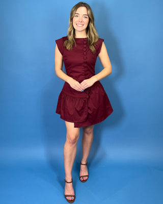 Rich burgundy sleeveless mini dress with a fitted bodice, statement covered buttons, and an asymmetrical drop-waist ruffle skirt.