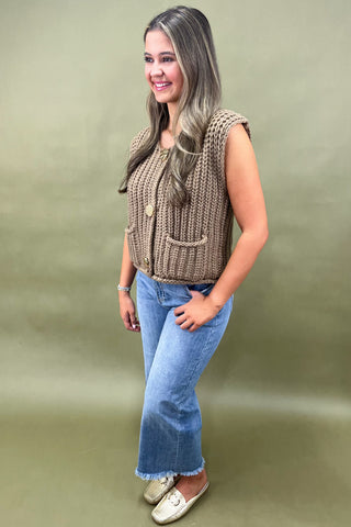 Woman wearing a brown knitted vest and blue jeans against a green background