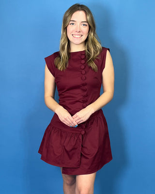 Rich burgundy sleeveless mini dress with a fitted bodice, statement covered buttons, and an asymmetrical drop-waist ruffle skirt.