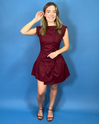Rich burgundy sleeveless mini dress with a fitted bodice, statement covered buttons, and an asymmetrical drop-waist ruffle skirt.