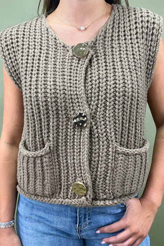 Person wearing a beige knitted vest with buttons on a plain background
