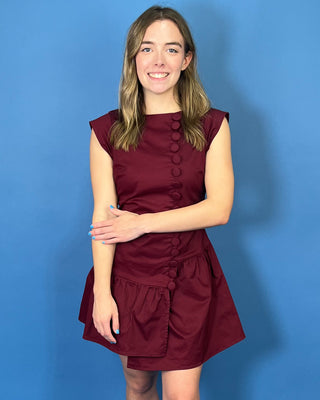 Rich burgundy sleeveless mini dress with a fitted bodice, statement covered buttons, and an asymmetrical drop-waist ruffle skirt.