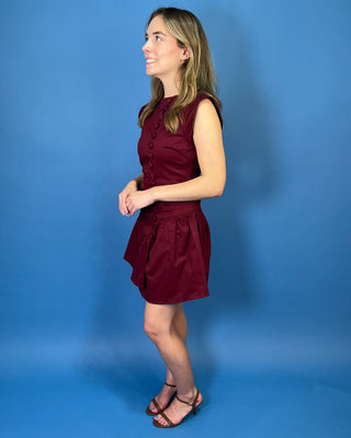 Rich burgundy sleeveless mini dress with a fitted bodice, statement covered buttons, and an asymmetrical drop-waist ruffle skirt.