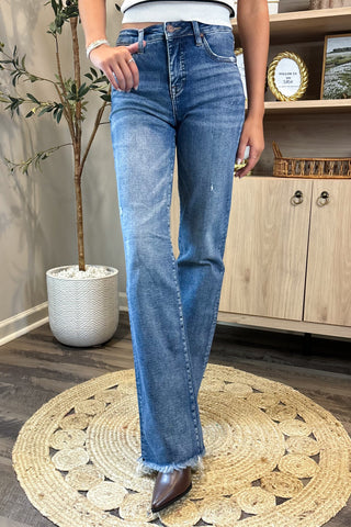 Person wearing blue jeans standing in a room with a plant and wooden cabinet.