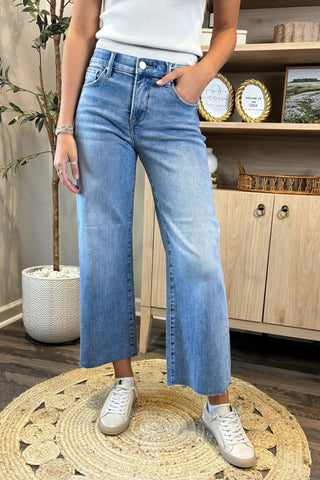 Person wearing blue jeans and white sneakers in a room with wooden flooring and a plant.