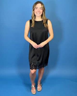 Black sleeveless satin shift dress with a classic round neckline, relaxed silhouette, and delicate lace trim along the hem.