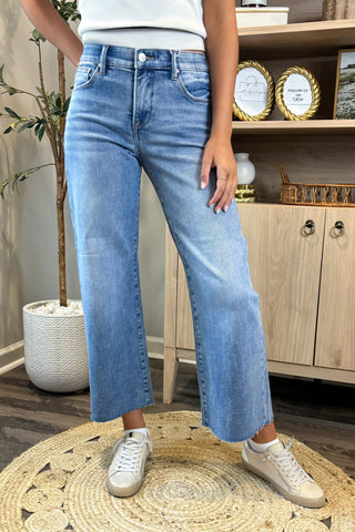 Person wearing light blue jeans and white sneakers in a home setting with a wooden cabinet and decorative items.