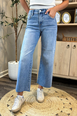 Person wearing light blue jeans and white sneakers in a room with a plant and wooden cabinet.