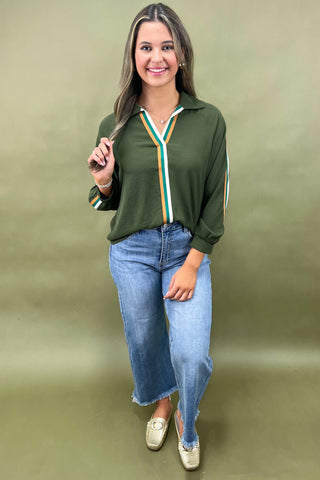 Woman wearing a green long-sleeve top with a striped collar and blue jeans on an olive green background
