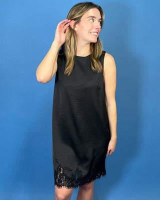 Black sleeveless satin shift dress with a classic round neckline, relaxed silhouette, and delicate lace trim along the hem.