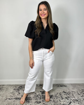 Model wearing a black puff sleeve blouse with a split neckline, styled with high-waisted white wide leg pants and clear slide sandals.