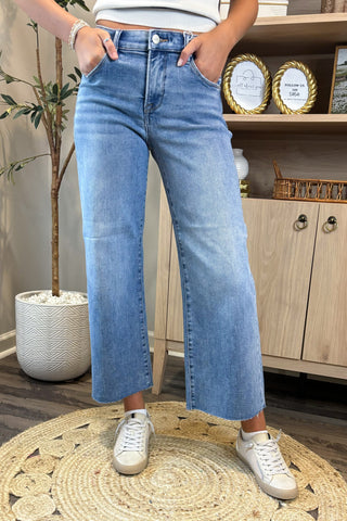 Person wearing blue jeans and white sneakers in a room with a plant and wooden cabinet.