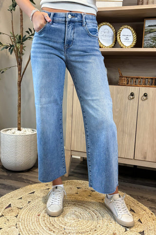 Person wearing blue jeans and white sneakers standing in a room with a plant and wooden cabinet.