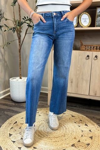 Person wearing blue jeans and white sneakers standing on a wooden floor with a plant and cabinet in the background.