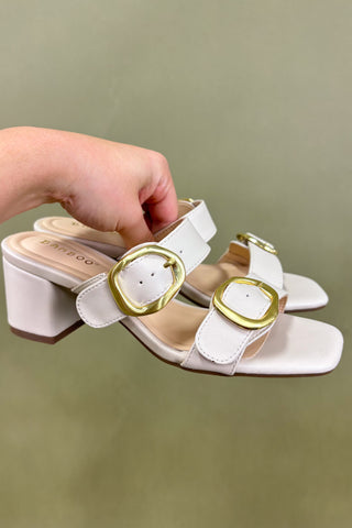 White sandals with gold buckles held by a hand against a green background